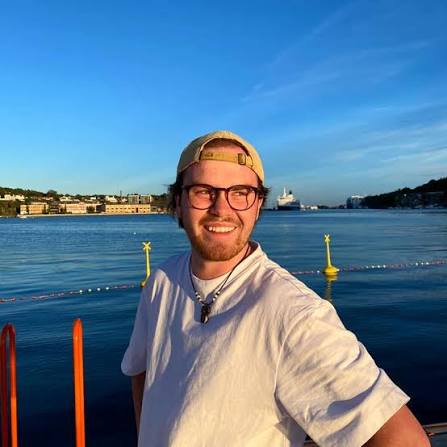 Odin Jean Aadland ute ved sjøkanten, med caps bak fram, briller og hvit t-skjorte